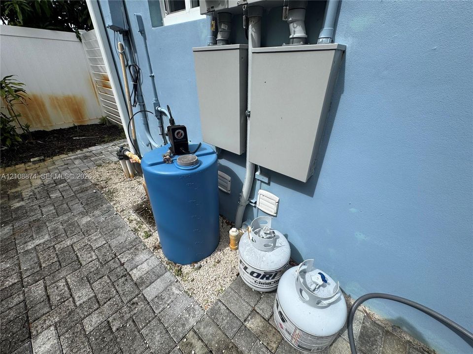 Exterior utility area featuring a blue water storage tank, two propane cylinders, two gray electrical disconnect enclosures, and stone paver ground cover