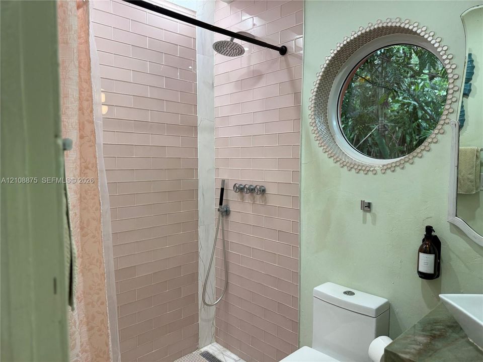 Shower featuring rose-toned rectangular wall tile, rain-style showerhead, and wall-mounted metal plumbing fixtures, complemented by a circular window with decorative trim and a vessel sink with stone-finish countertop