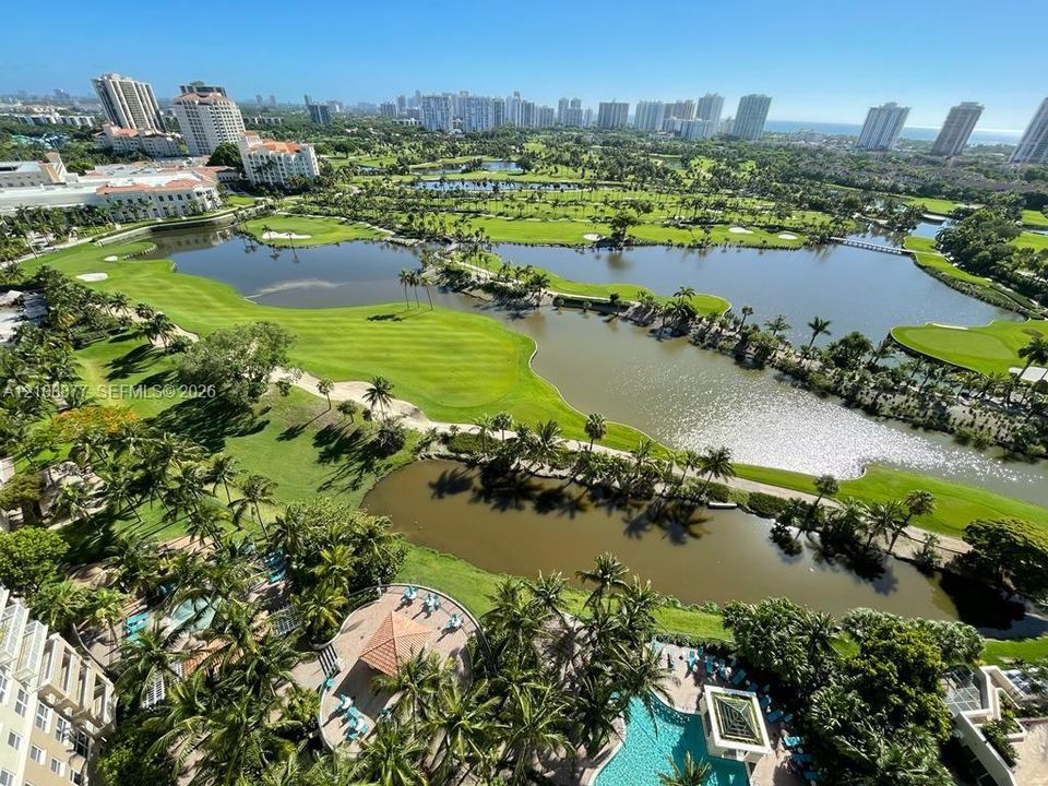 Aerial perspective featuring a large swimming pool, paved pool deck, and a pavilion structure, overlooking expansive green space, multiple water features, and a skyline horizon