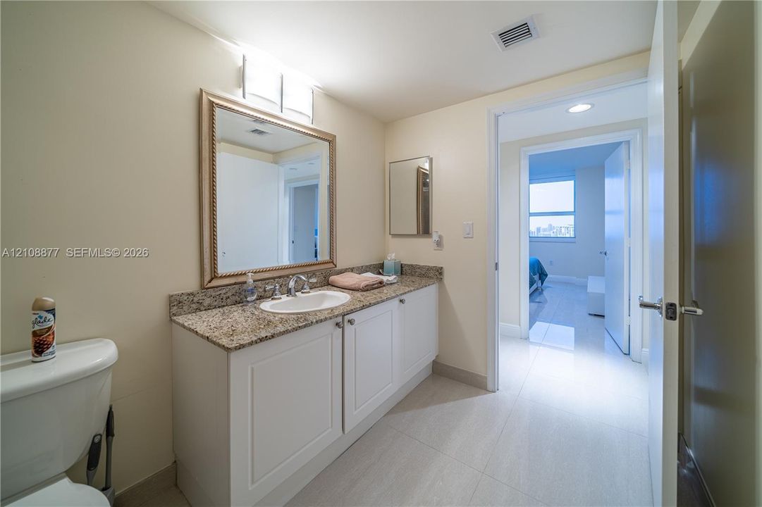 Bathroom featuring a stone-top vanity with an integrated oval sink, white shaker-style cabinetry, a framed wall mirror, and large-format tile flooring