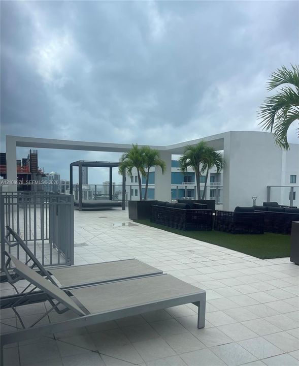 Rooftop terrace featuring large-format tile flooring, a minimalist white pergola structure, glass panel perimeter railing, and integrated artificial turf area