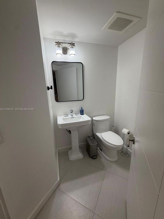 Pedestal sink, rectangular wall-mounted mirror with a dark frame, two-light fixture, and light-toned tile flooring