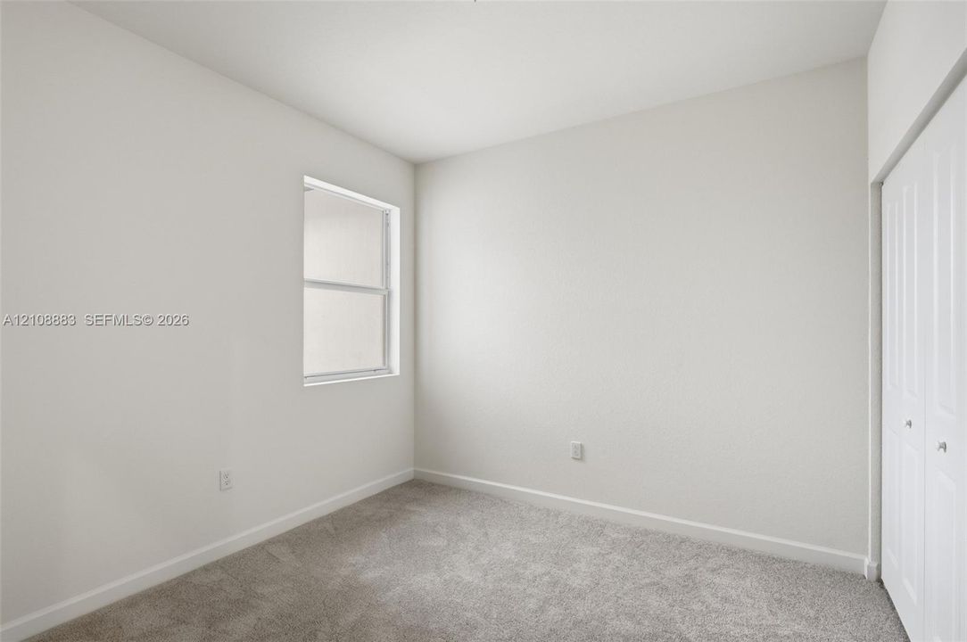 Neutral-toned interior space featuring light-colored wall paint, carpeted flooring, a single window with white trim, and white bi-fold closet doors