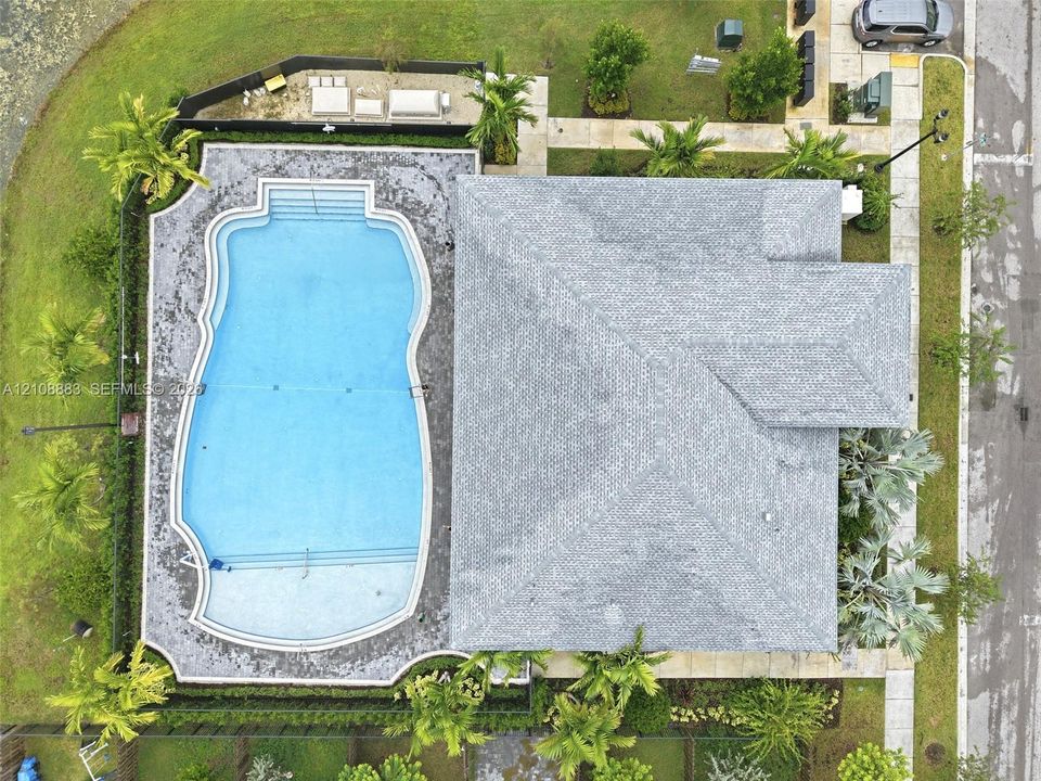 Aerial perspective featuring a custom-shaped swimming pool with tiered entry steps, expansive paver decking, mature perimeter landscaping, and a multi-faceted shingle roof