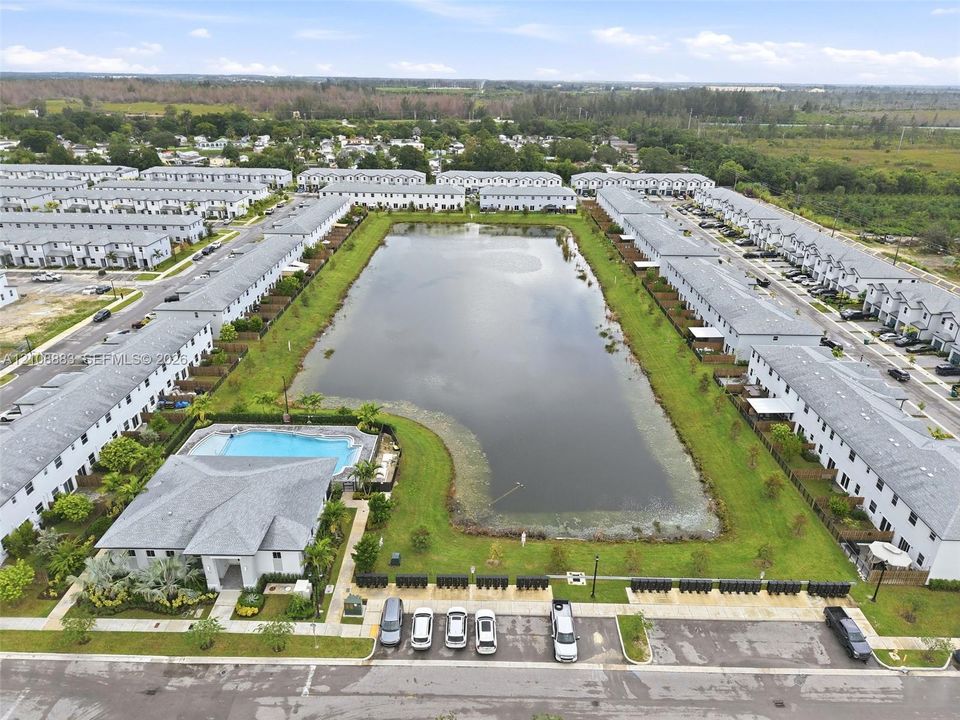 Aerial perspective of a community featuring a central retention pond, paved parking areas, and a communal swimming pool with surrounding deck