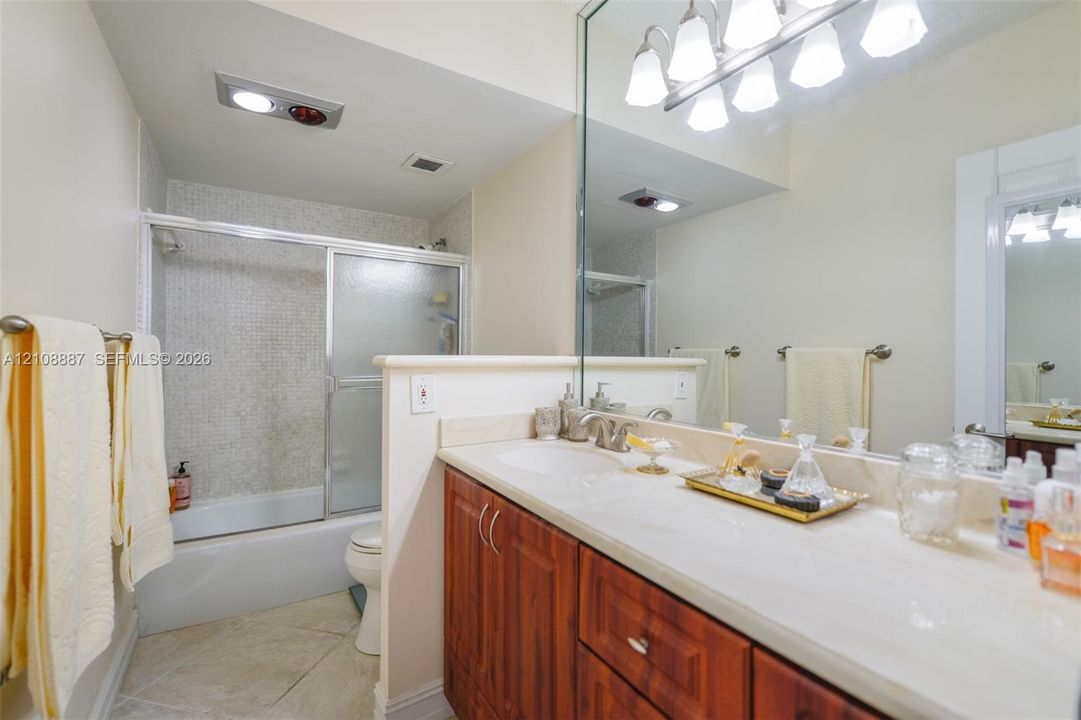 Bathroom featuring a solid-surface countertop, wood-finish cabinetry, a full-width vanity mirror, a combination tub and shower with a sliding glass enclosure, and tile flooring