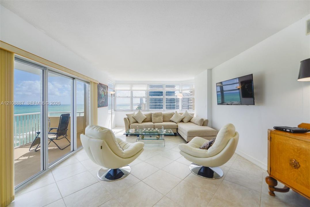 Living area featuring neutral tile flooring, expansive floor-to-ceiling glass sliding doors, wrap-around window configurations, and a private balcony with ocean frontage
