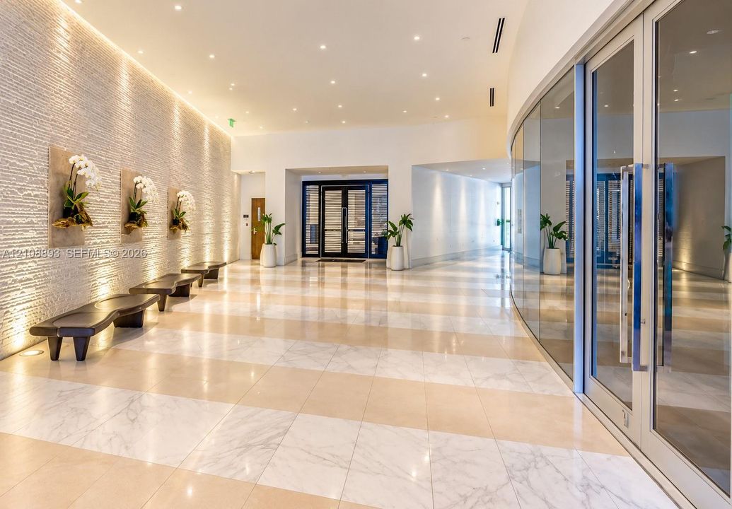 Grand lobby featuring a textured stone accent wall, large-format tile flooring, recessed ceiling lighting, and a curved floor-to-ceiling glass wall enclosure