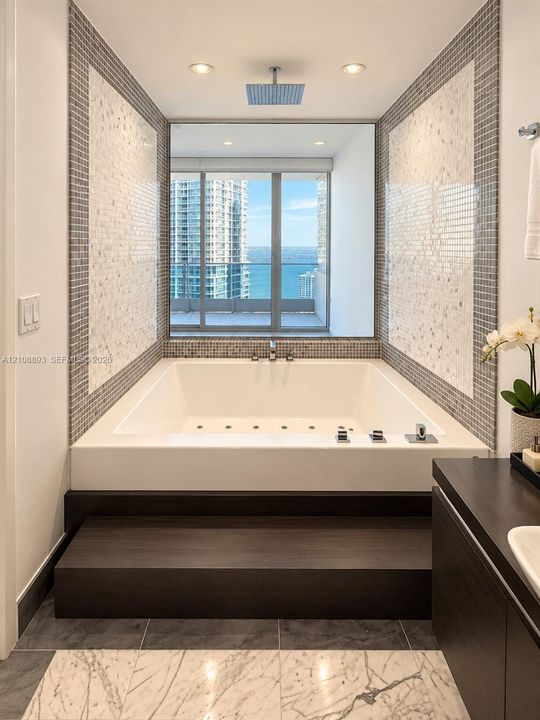 Built-in soaking tub with a raised dark wood-finish platform, overhead rainfall showerhead, mosaic tile surround, and expansive window showcasing water and skyline scenery