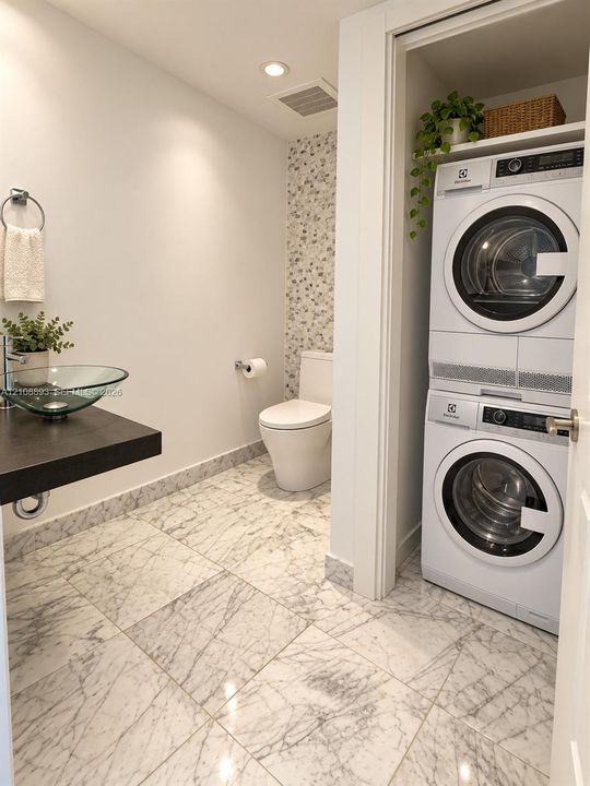 Full-bathroom featuring large-format stone-patterned floor tile, glass vessel sink with dark floating vanity, integrated laundry closet, and decorative stone-mosaic wall accent