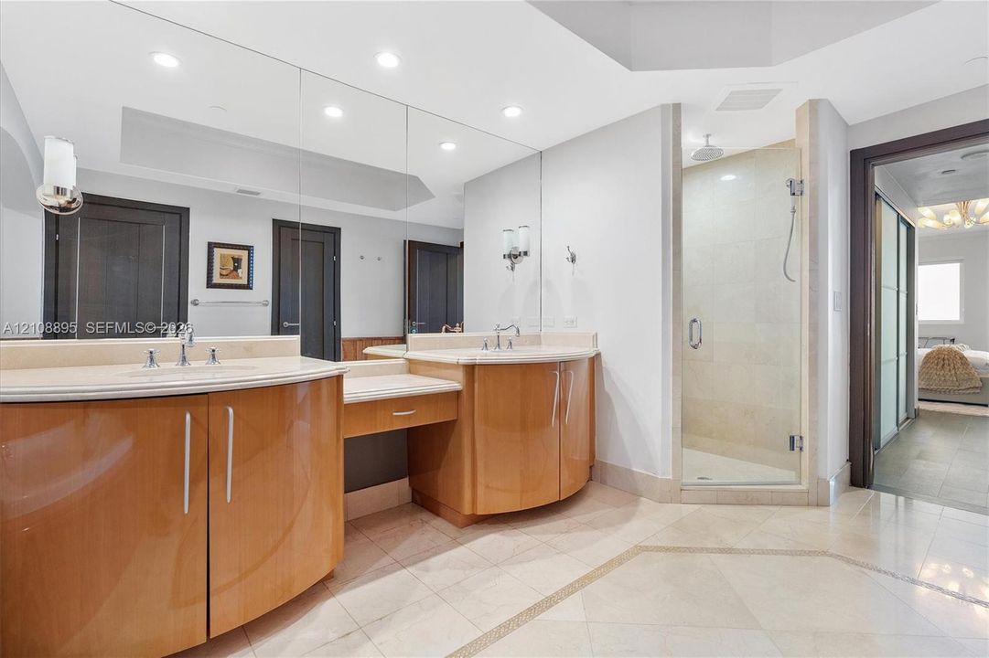 Curved wood-finish cabinetry, neutral stone-surfaced countertops with integrated sinks, full-width wall mirror, and a glass-enclosed shower with wall-mounted fixture