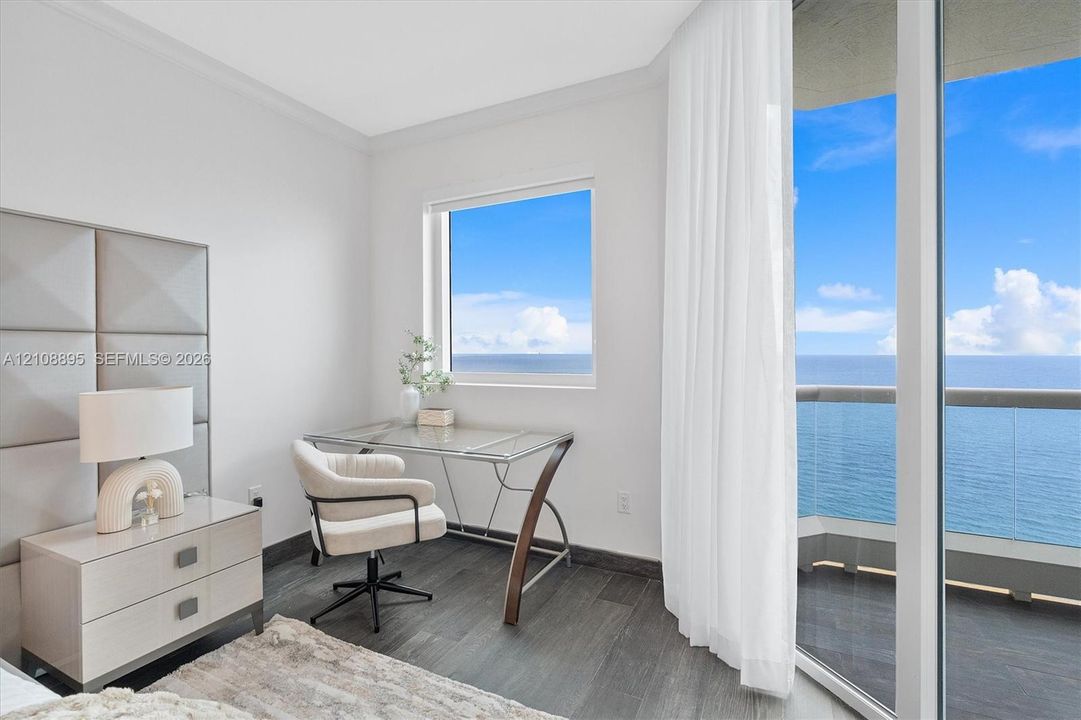 Floor-to-ceiling glass sliding doors opening to a balcony with ocean scenery, wood-finish flooring, and a multi-panel upholstered wall accent