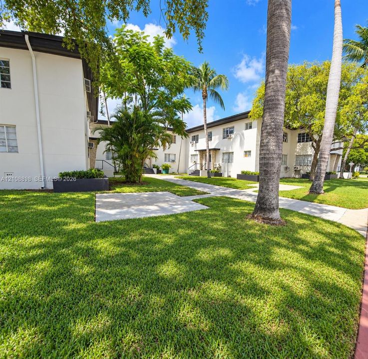 Stucco exterior architecture featuring multi-story building structures, concrete walkways, manicured lawn areas, mature palm trees, and dark-finished rectangular planter boxes