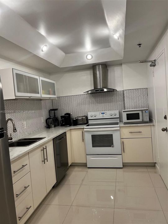 Kitchen featuring high-gloss cabinetry, stone-finish countertops, herringbone-patterned tile backsplash, stainless steel chimney-style range hood, and recessed tray ceiling lighting