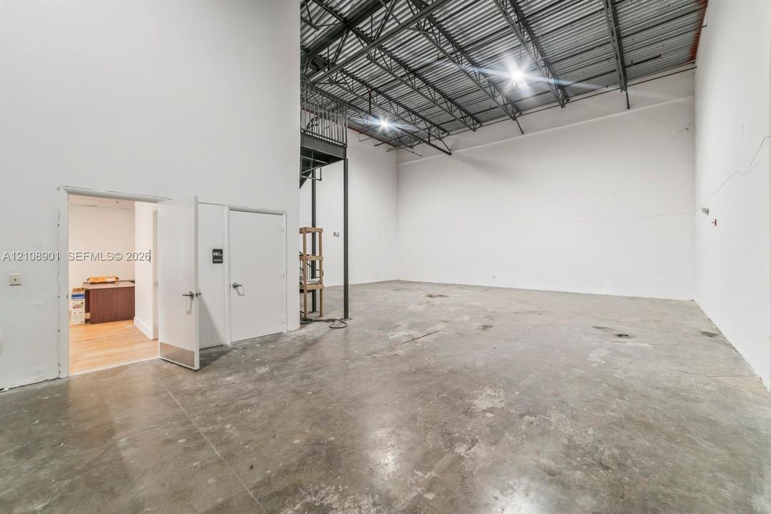Open-concept warehouse space featuring high ceilings with exposed structural steel, durable concrete flooring, white interior walls, and an integrated metal mezzanine structure