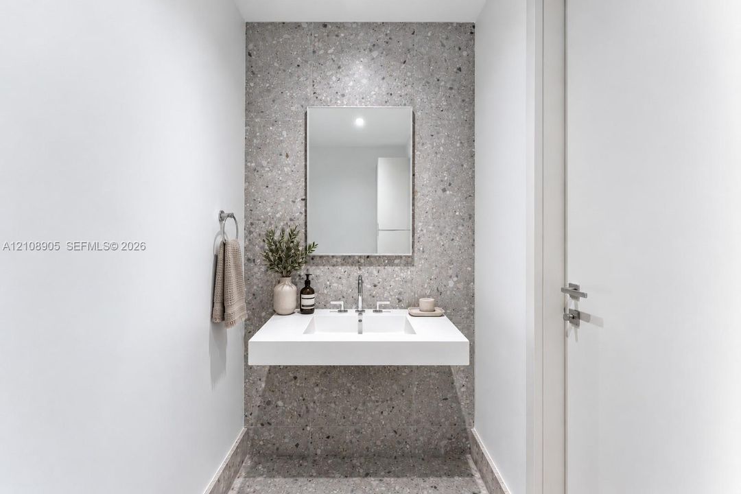 Floating vanity with a rectangular integrated sink, polished metal widespread faucet, rectangular wall-mounted mirror, and speckled stone-finish wall cladding