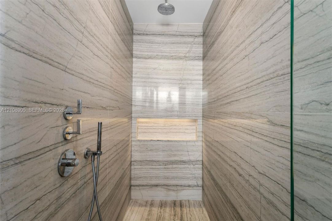 Walk-in shower featuring floor-to-ceiling stone-tiled walls, a built-in recessed niche, chrome wall-mounted fixtures, a handheld sprayer, and a ceiling-mounted rainfall showerhead