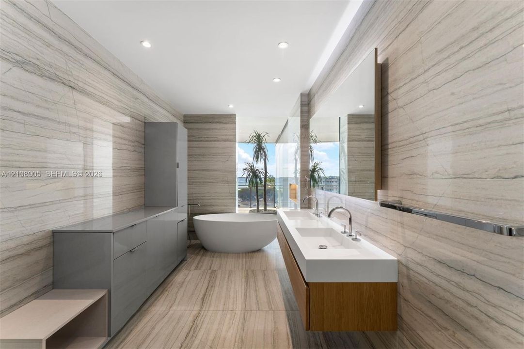 Full-height stone wall cladding, floating wood-finish dual vanity with integrated sinks, freestanding soaking tub, expansive wall-mounted mirror, and sleek grey cabinetry
