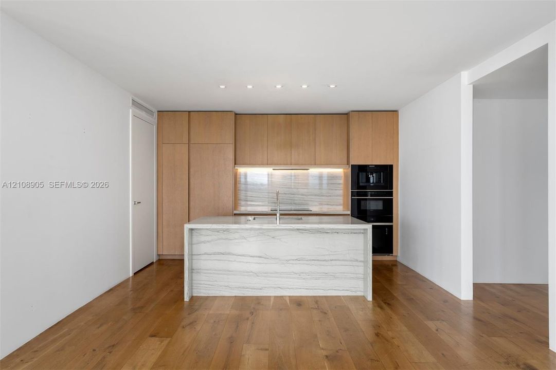 Modern kitchen featuring custom wood-finish cabinetry, a waterfall-edge stone island, built-in dual ovens, integrated cooktop, and wood-finish flooring