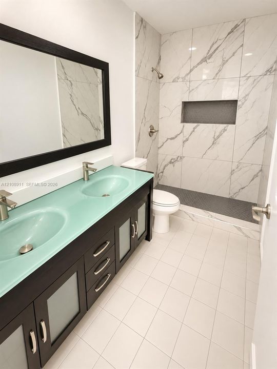 Double vanity featuring a frosted glass countertop, dual undermount sinks, espresso-finished cabinetry with glass inserts, and a walk-in shower with large-format stone-patterned wall tile and an integrated recessed niche