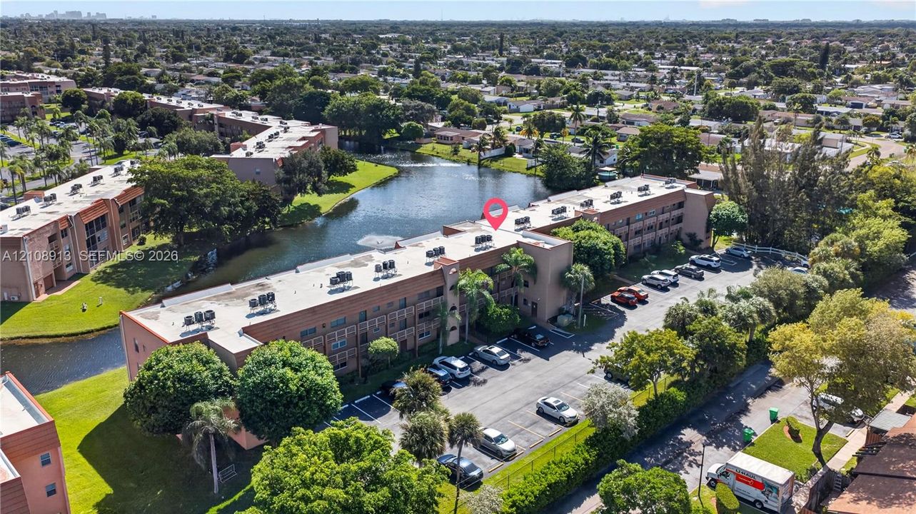 Aerial perspective of a multi-story residential building featuring water frontage, an on-site surface parking lot, and mature landscaping