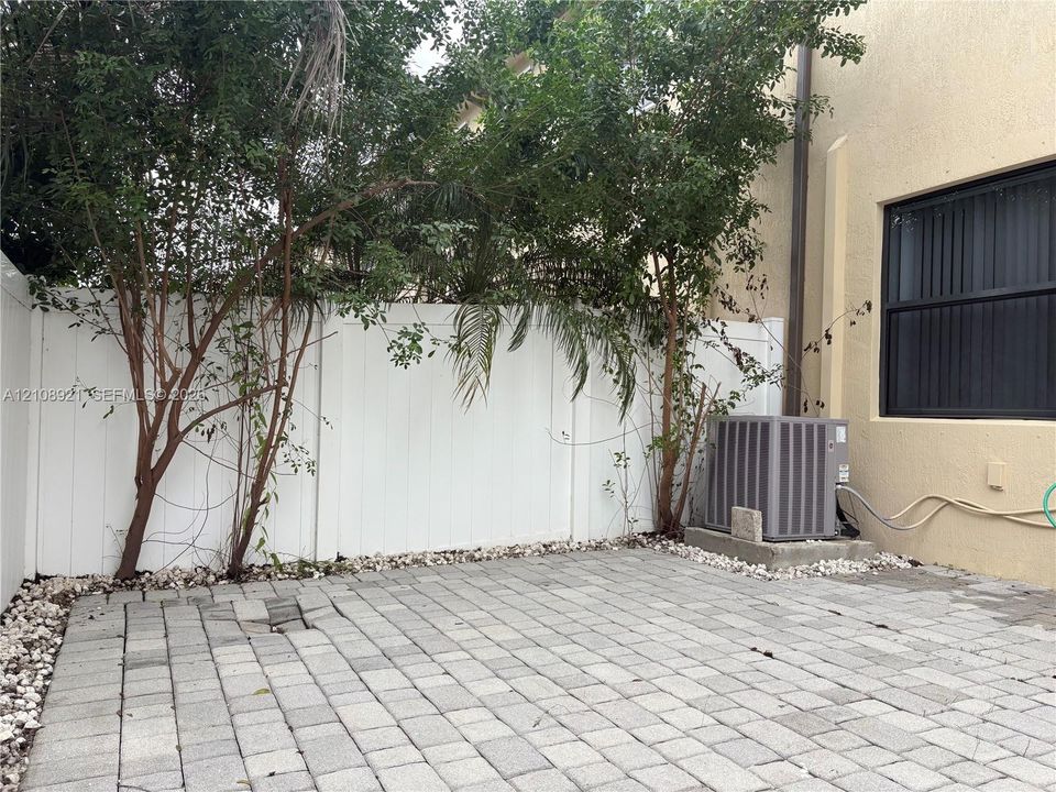Paved stone patio featuring a white vinyl privacy fence, established trees, and exterior HVAC unit