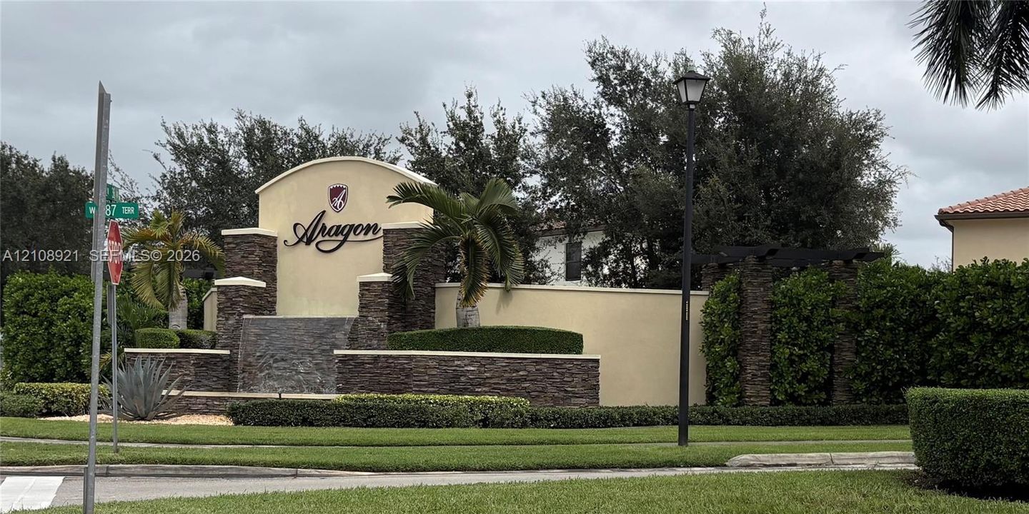Community entrance featuring a stone-accented monument wall, integrated water feature, mature landscaping, and an arched architectural header
