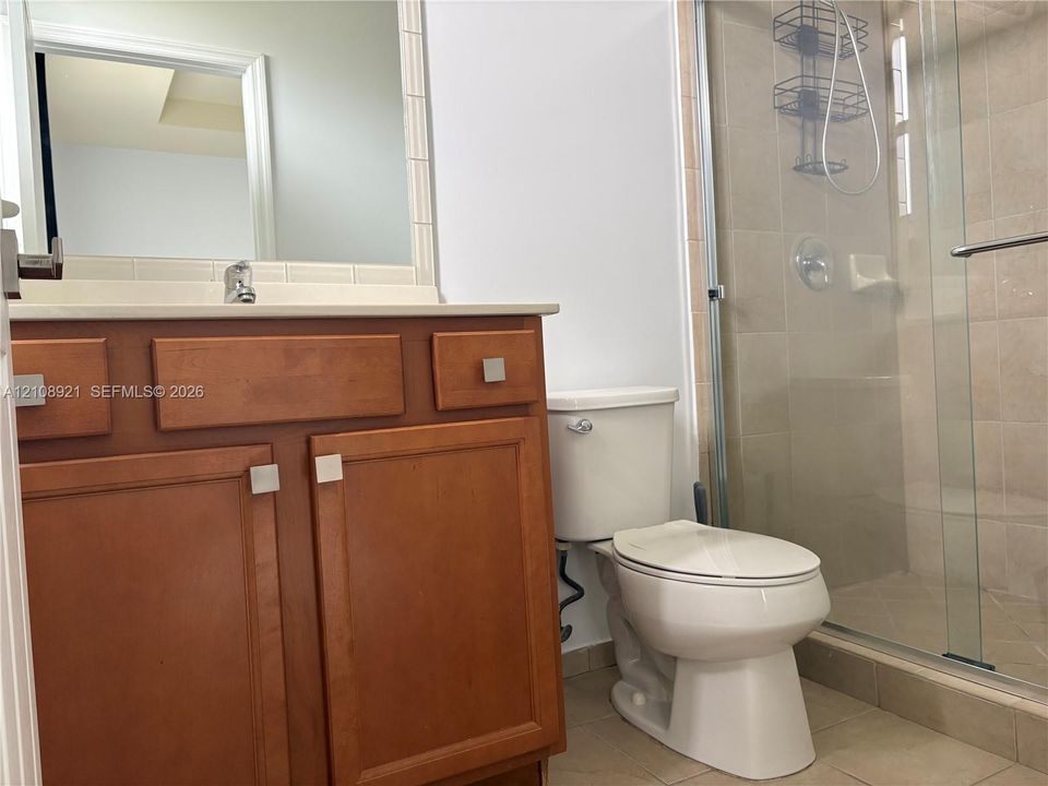 Wood-finish vanity cabinet, solid-surface countertop, frameless mirror, and glass-enclosed shower with tile surround