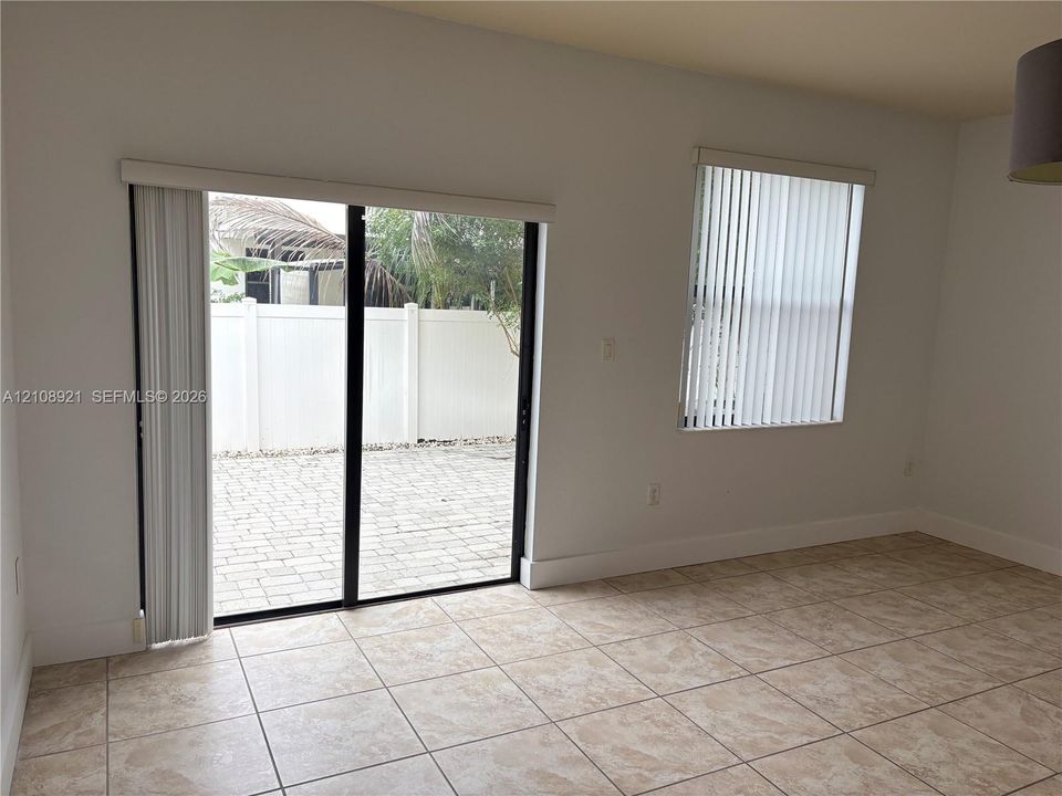 Interior space featuring neutral-toned tile flooring, sliding glass door with vertical blinds, single-pane window with vertical blinds, and a paved exterior patio area enclosed by a privacy fence