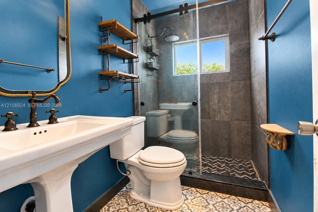 Bathroom featuring a white pedestal sink with oil-rubbed bronze fixtures, a walk-in shower with charcoal large-format tile walls, a sliding glass door, patterned floor tile, and built-in industrial-style shelving