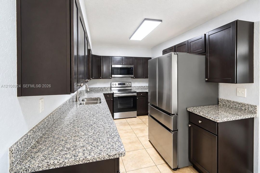 Kitchen featuring dark wood-finish cabinetry, stone-finish countertops, stainless steel appliances, a double-basin undermount sink, and tile flooring