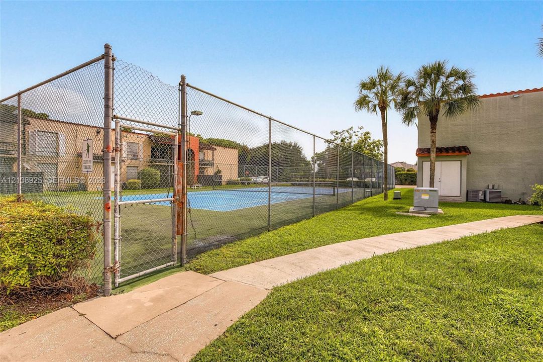 Tennis court enclosed by chain-link fencing, accompanied by a paved walkway and manicured grounds