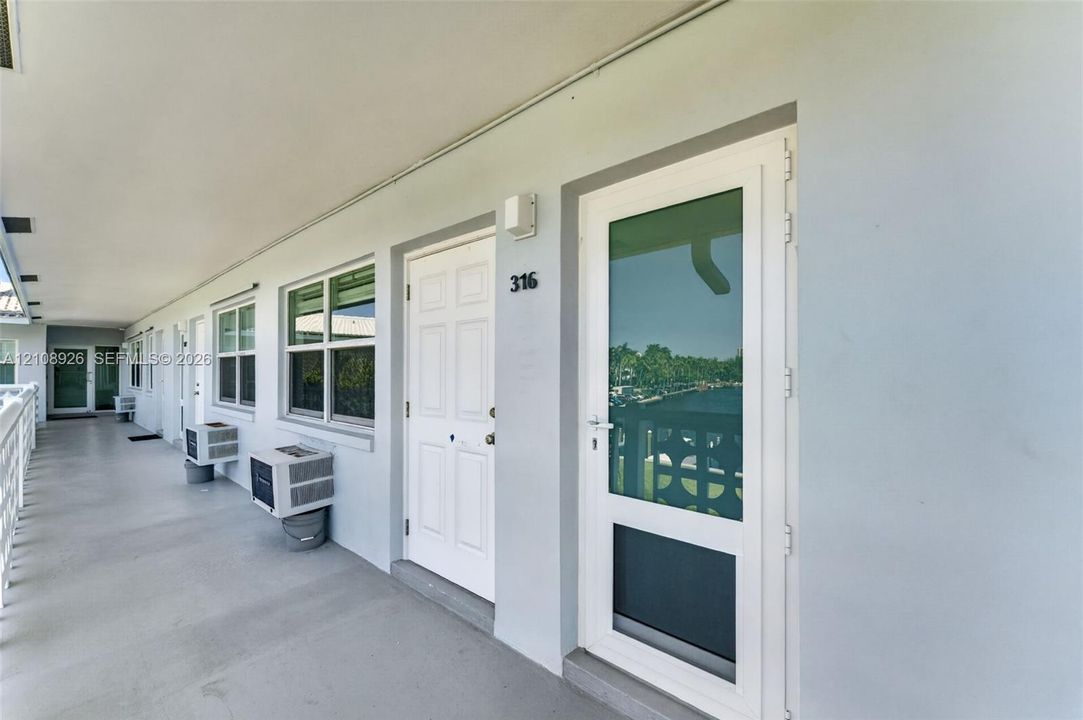 Covered exterior walkway featuring a six-panel entry door, glass-paneled storm door, dual-pane windows, and exterior-mounted cooling units