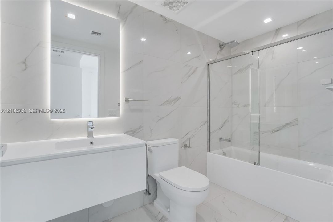 Bathroom featuring large-format stone-patterned wall and floor tile, a modern floating vanity with an integrated sink, a frameless glass tub enclosure, and an LED-backlit mirror
