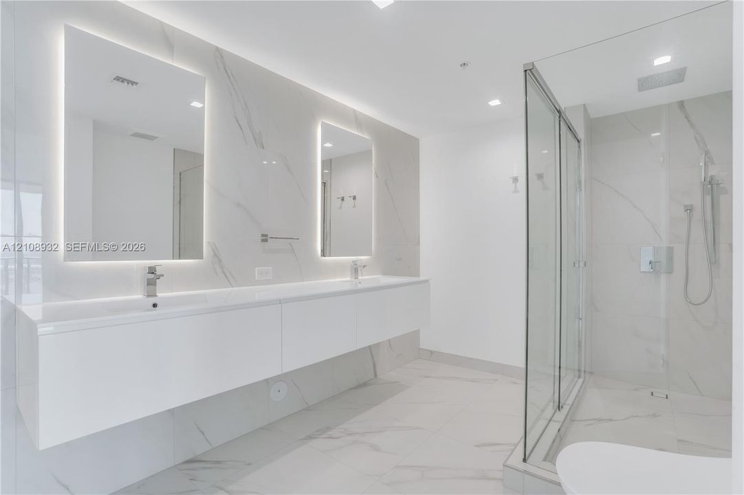 Floating dual vanity with integrated sinks, back-lit mirrors, walk-in glass shower enclosure, and full-height stone-finish wall tiling