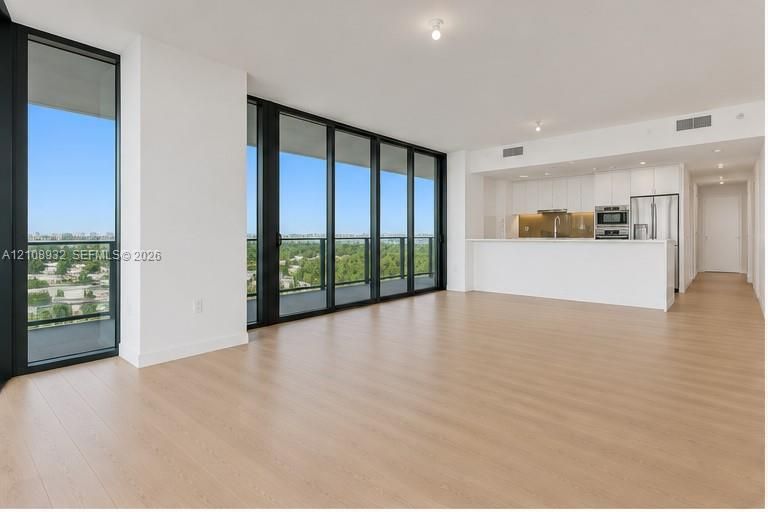 Open-concept living space featuring floor-to-ceiling windows with dark frames, wood-finish flooring, and a kitchen equipped with white flat-panel cabinetry and stainless steel appliances