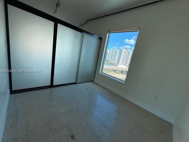 Polished concrete flooring, floor-to-ceiling sliding closet doors with frosted glass panels and black frames, exposed industrial piping, and a large window featuring an urban skyline