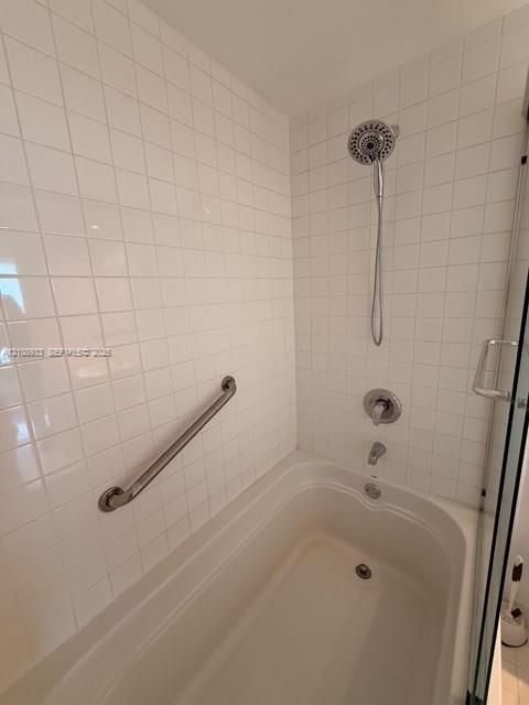 Bathtub and shower combination featuring white square tile surround, chrome hand-held shower fixture, single-handle pressure balance valve, and mounted metal grab bar