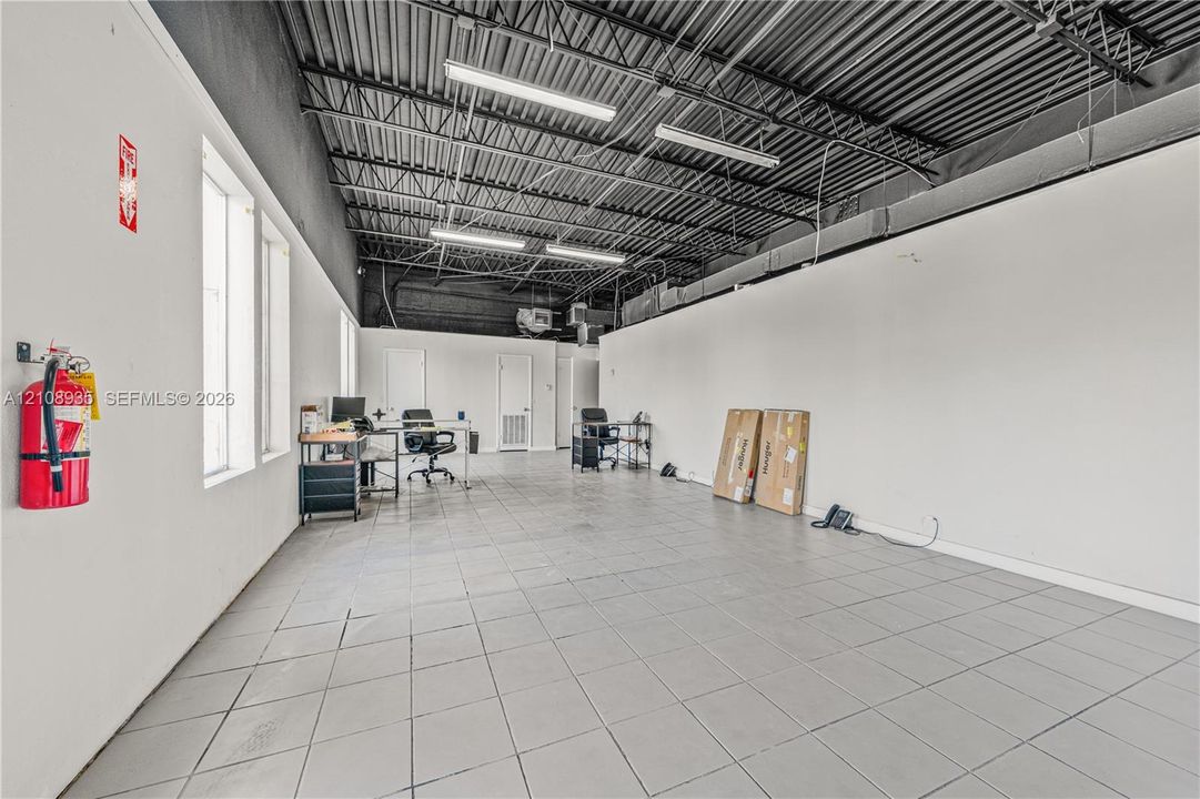 Expansive interior featuring high, exposed-joist ceilings, industrial-style black painted rafters, large rectangular ceramic tile flooring, and linear fluorescent lighting fixtures