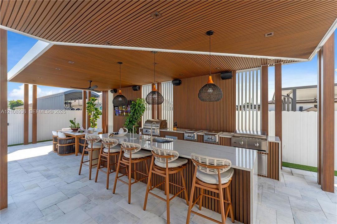 Covered outdoor kitchen featuring a slatted wood ceiling, large stone-topped island with integrated sink, stainless steel built-in grill stations, recessed lighting, and large woven pendant fixtures