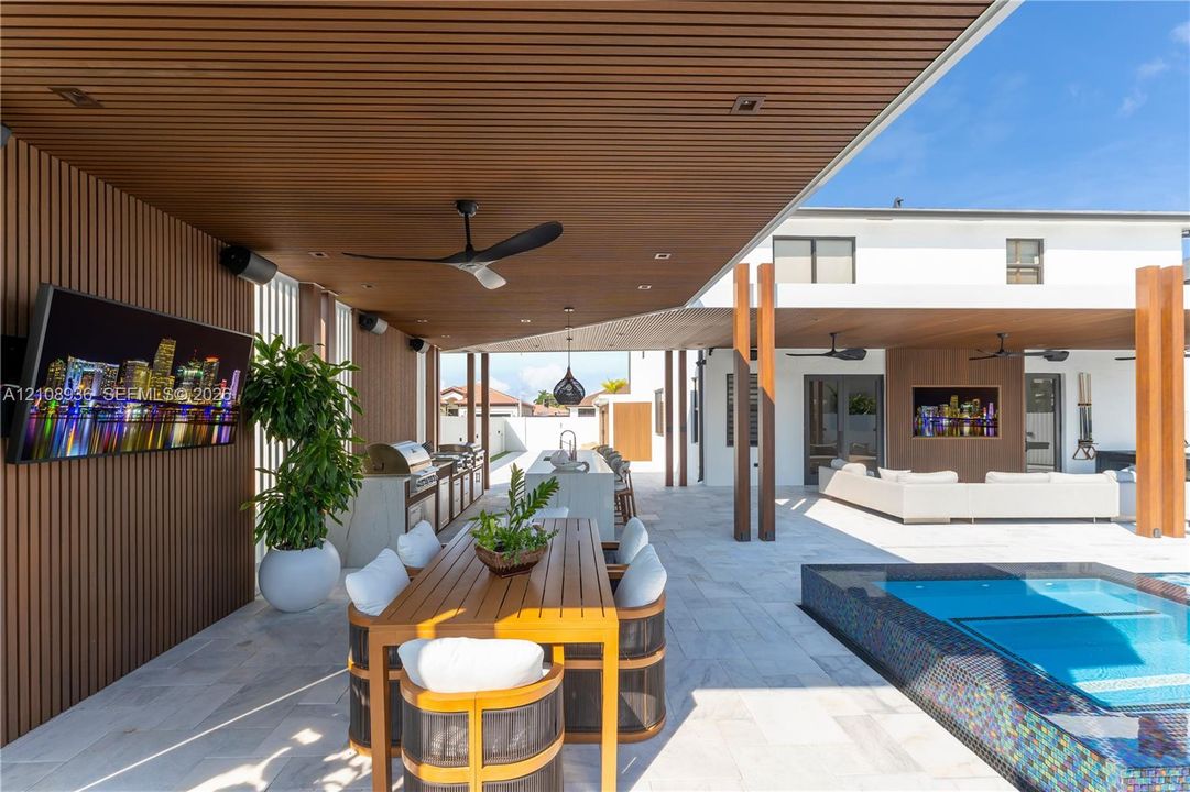 Covered outdoor patio featuring a slatted wood-finish ceiling, stone flooring, built-in kitchen island, recessed lighting, and a private pool with tiled surround