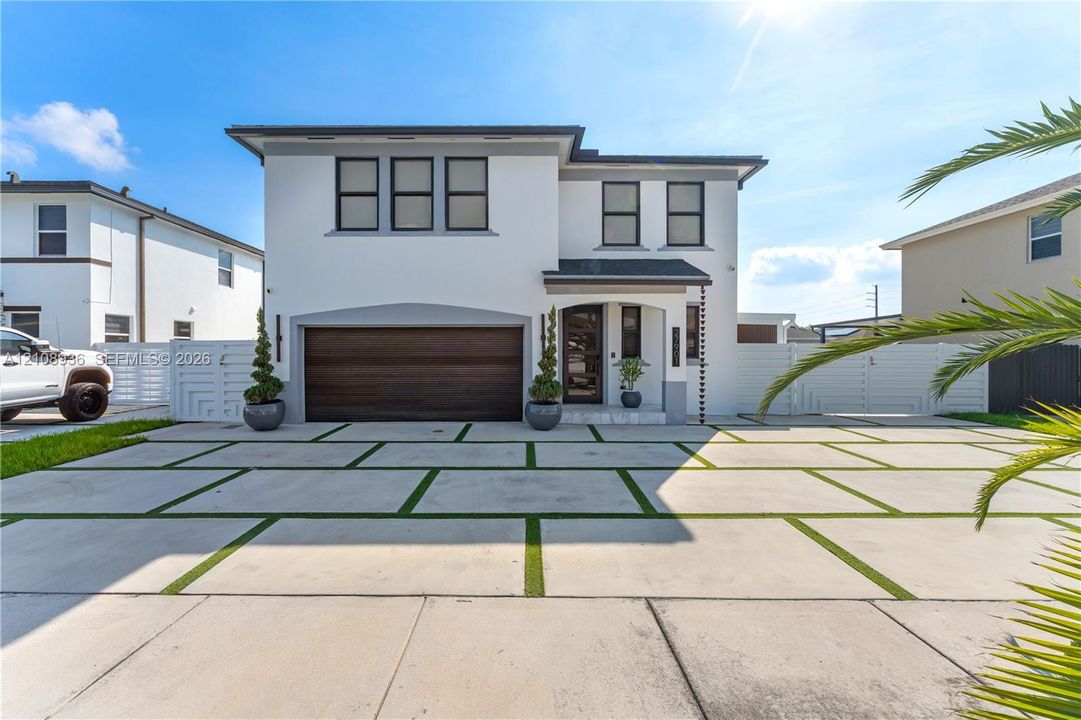 Contemporary two-story exterior featuring a smooth white stucco finish, a dark wood-tone garage door, grid-patterned concrete paver driveway with integrated turf, and matching perimeter fencing