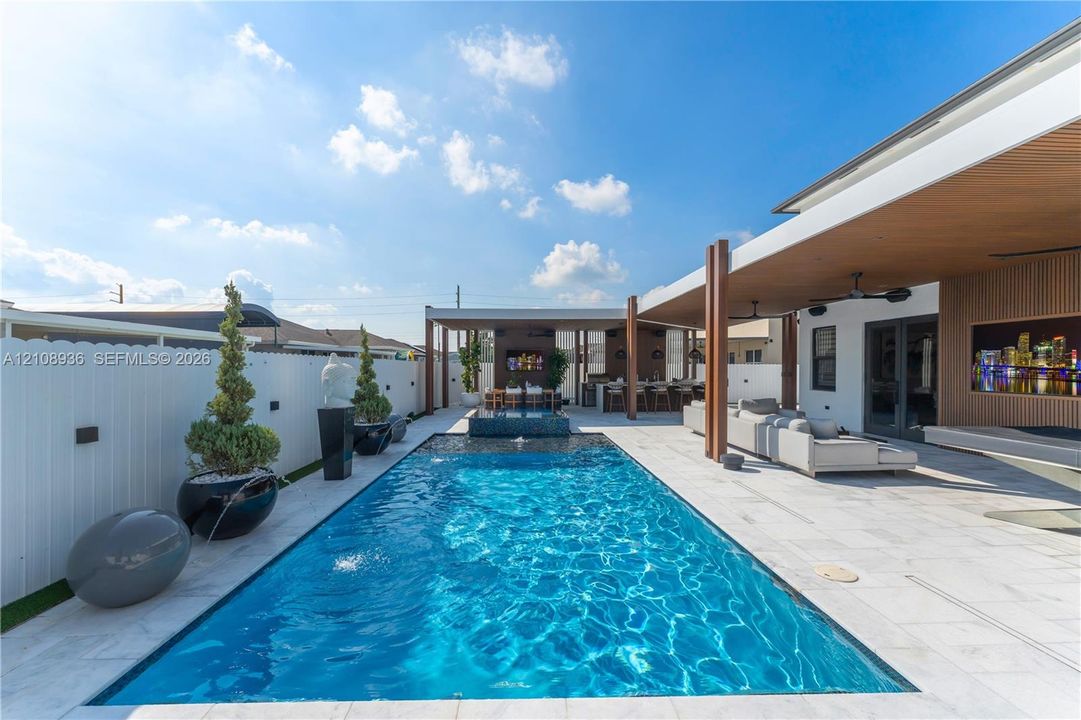 Rectangular swimming pool featuring a tiled deck, integrated water feature, covered patio with wood-finished ceiling accents, and attached outdoor kitchen area