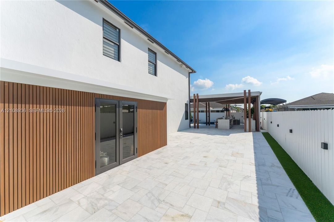 Exterior white stucco facade featuring wood-slat siding, double glass entry doors, expansive stone paver patio, and a covered pergola with a built-in outdoor kitchen