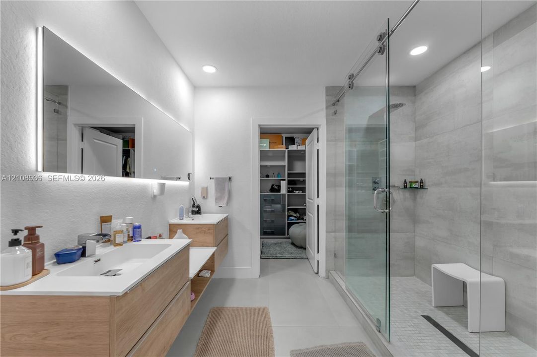 Floating dual vanity with wood-finish cabinetry, frameless glass walk-in shower featuring large-format tile surround, recessed lighting, and integrated LED-lit vanity mirror