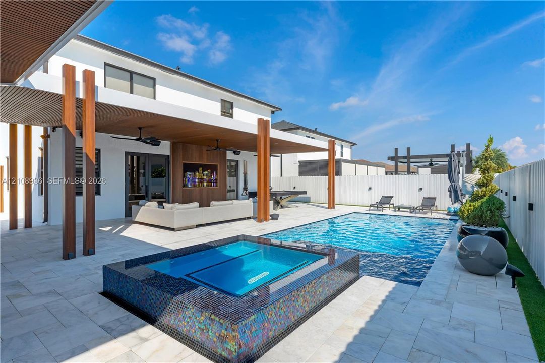 Covered outdoor patio with wood-paneled ceiling, integrated ceiling fans, in-ground pool, spa with iridescent tile finish, and light-toned paver hardscaping