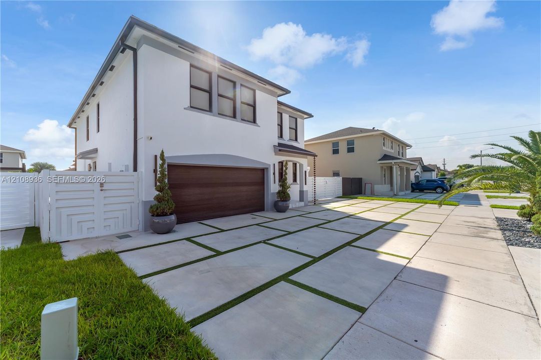 Two-story stucco exterior featuring a wood-finish garage door, large-format concrete paver driveway with turf accents, and a white privacy gate