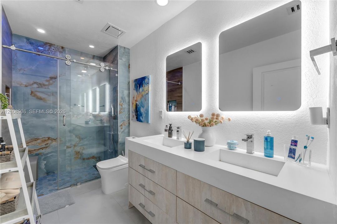 Dual-sink vanity with light-tone cabinetry and white countertop, frameless glass shower enclosure featuring blue stone-style tile surround, and dual wall-mounted mirrors with integrated LED backlighting