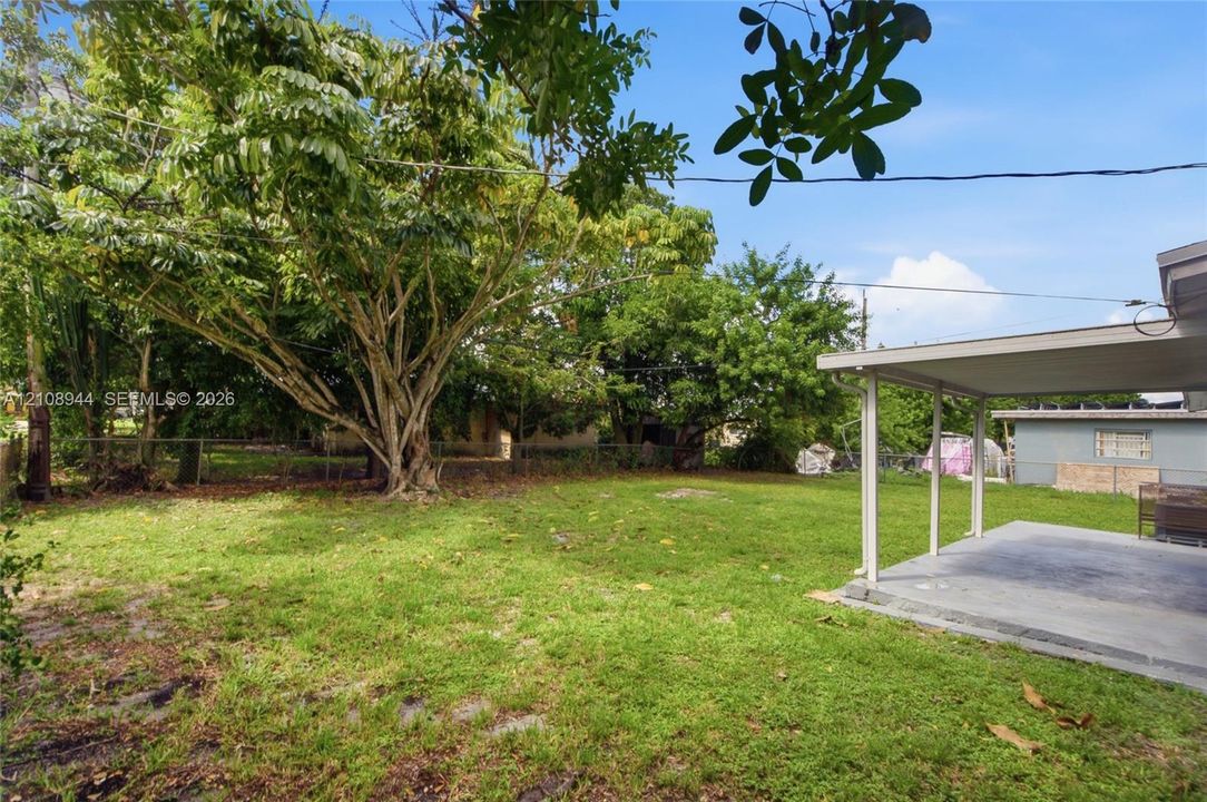 Backyard featuring a concrete patio, covered porch, mature trees, chain-link fencing, and grass lawn