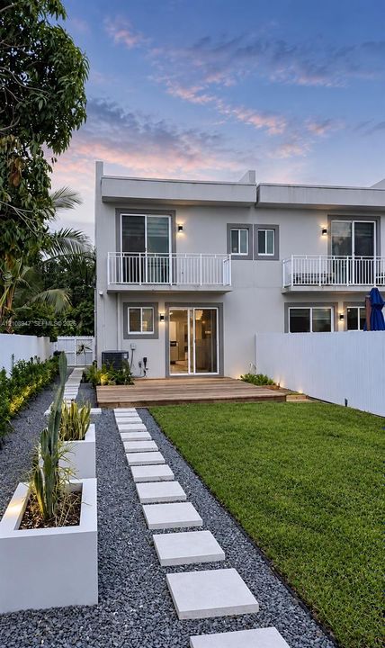 Exterior facade featuring second-story balconies, rear wood decking, manicured lawn, stone paver pathway, and geometric concrete planters